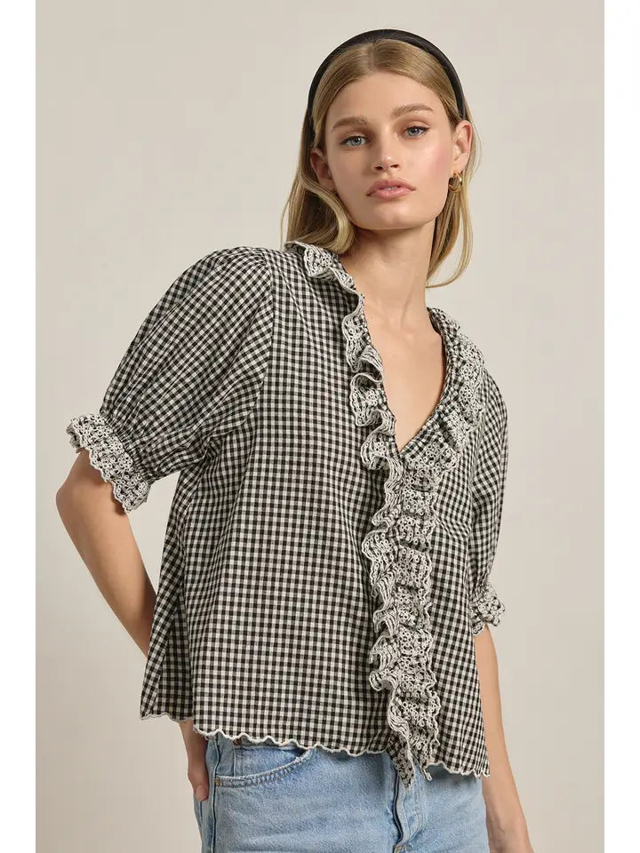 A woman wears the Zertz GINGHAM LACE RUFFLE BLOUSE, featuring black and white gingham with lace ruffles and scalloped edges, paired with light blue jeans. She has straight blonde hair, a black headband, and a neutral expression.