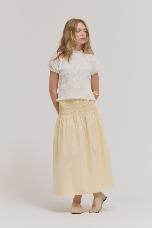 A woman with light brown hair and beige shoes stands against a plain background, wearing a white textured top and the Zertz SMOCKED WAIST SKIRT in light yellow, an effortless essential that flatters with its high smocked waist. Her hands are behind her back.