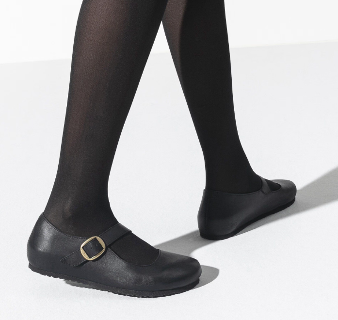 A person wearing black tights and Zertz TRACY LEATHER BLACK 4 Mary Jane shoes with a gold buckle walks on a light surface, highlighting a feminine aesthetic with only the lower legs and feet visible.