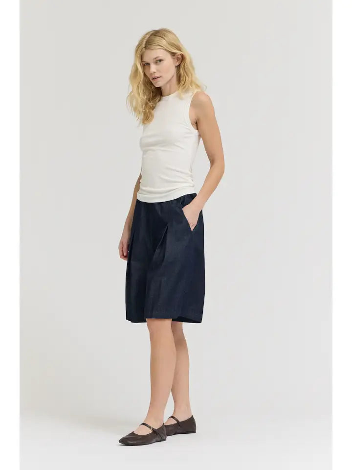 A woman with blonde hair and a relaxed posture stands against a light background, wearing the Zertz SHEER TANK TOP, dark knee-length shorts with pockets, and brown flat shoes.