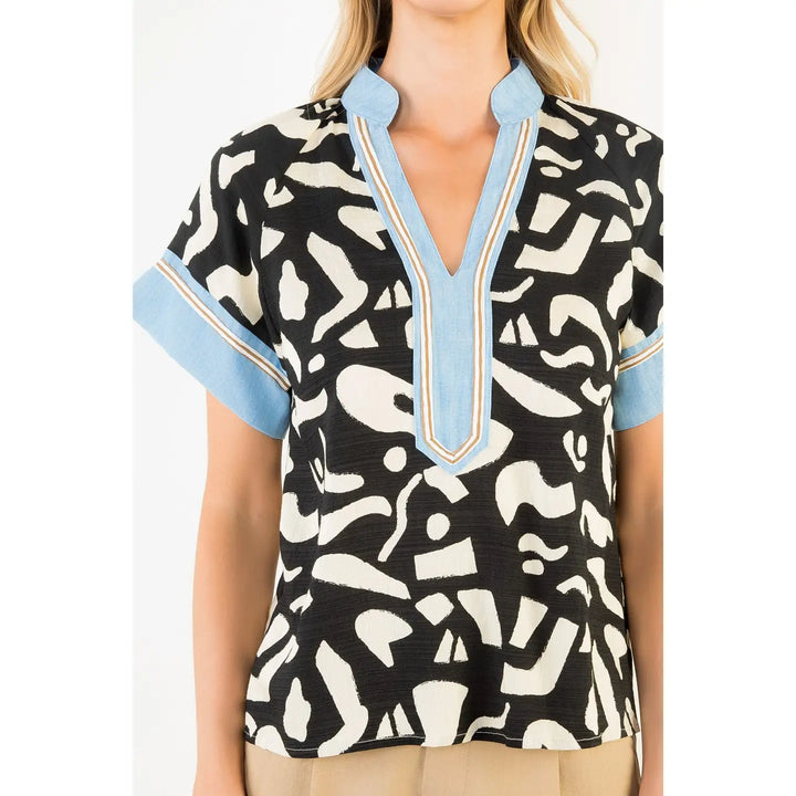 A woman wears the Zertz ABSTRACT PRINT TOP, a modern black short-sleeve top with white abstract shapes, light blue cuffs, and a blue V-neckline trimmed in gold. The photo shows her from shoulders to hips.