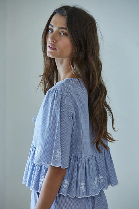 A woman with long, wavy brown hair looks over her shoulder wearing the Zertz CELESTINE WOVEN TOP, a blue and white striped blouse featuring a ruffled hem and breathable fabric against a softly lit, plain background.