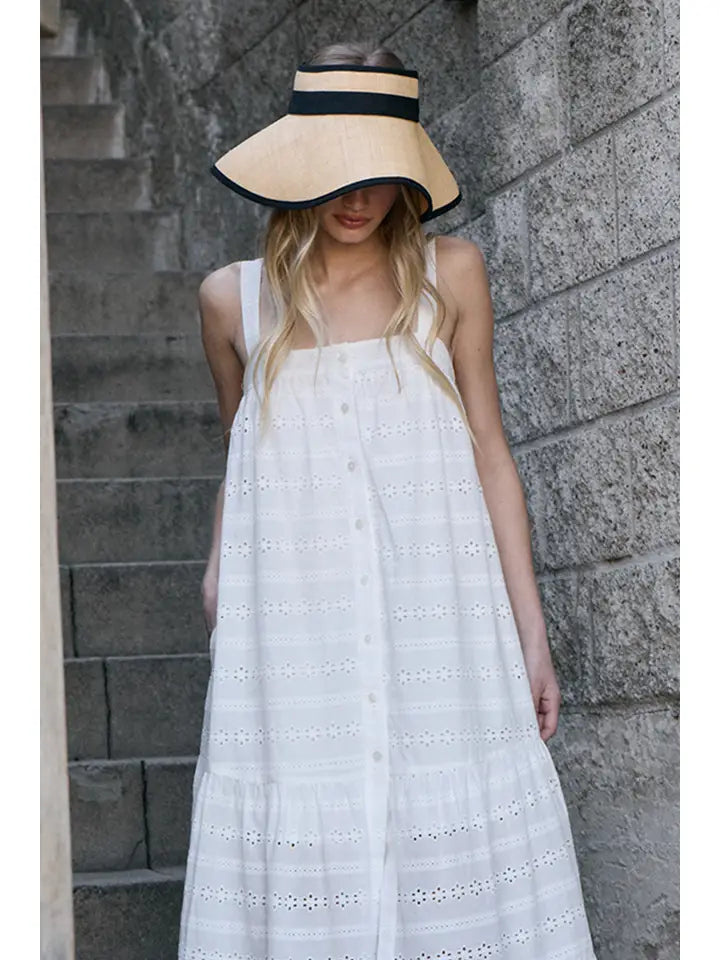 A woman wearing the Zertz EYELET BUTTON FRONT MAXI DRESS stands on stone stairs by a textured wall, accessorized with a wide-brimmed tan visor hat with a black ribbon. Her long blond hair is down as she looks downward.