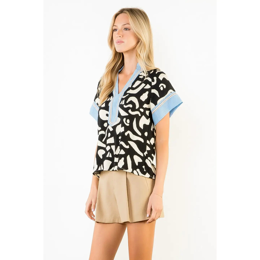 A woman models the Zertz ABSTRACT PRINT TOP—an abstract print blouse with blue cuffs and collar—against a white background, paired with beige shorts. Her long blonde hair and relaxed pose emphasize her modern, statement-making style.