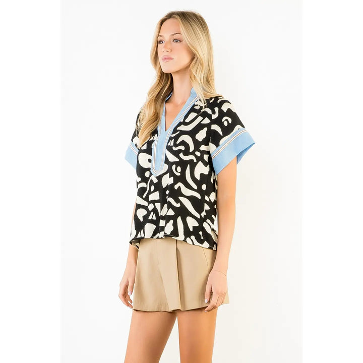 A woman models the Zertz ABSTRACT PRINT TOP—an abstract print blouse with blue cuffs and collar—against a white background, paired with beige shorts. Her long blonde hair and relaxed pose emphasize her modern, statement-making style.