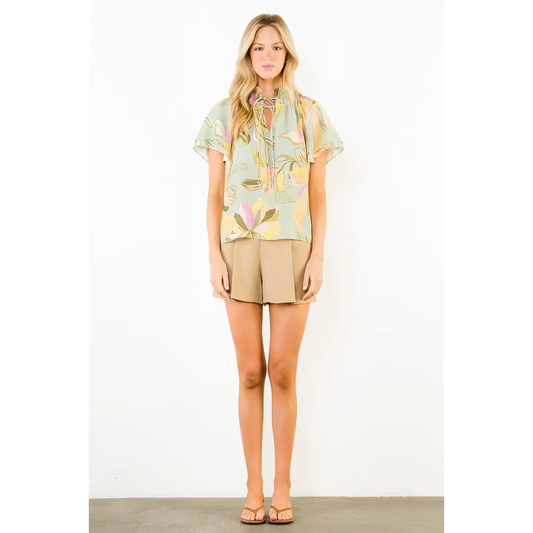 A woman with long blonde hair and a neutral expression wears the Zertz FLOWY FLOWER TOP WITH TIE FRONT, tan shorts, and beige sandals against a white background.