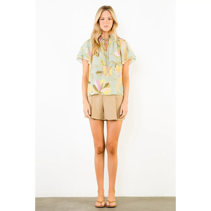 A woman with long blonde hair and a neutral expression wears the Zertz FLOWY FLOWER TOP WITH TIE FRONT, tan shorts, and beige sandals against a white background.
