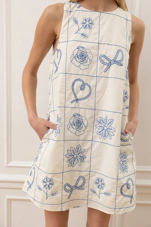 A woman wears the Zertz FLORAL PATTERN SLVLESS MINI DRESS, a cream, above-the-knee shift with blue embroidered florals, hearts, and ribbons in a grid design. The handcrafted dress features pockets and is sleeveless.