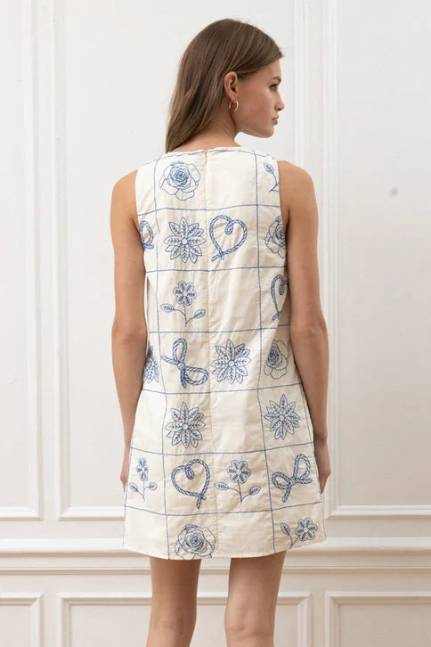 A woman with light brown hair models the Zertz FLORAL PATTERN SLVLESS MINI DRESS in cream, showcasing blue embroidered floral and ribbon grid patterns. She stands back to camera by a white paneled wall, highlighting its handcrafted details.