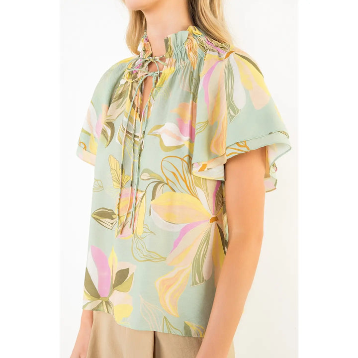 A woman wears the Zertz FLOWY FLOWER TOP WITH TIE FRONT in light green, featuring a pink, yellow, and beige floral pattern, short flutter sleeves, a ruffled tie-front neckline, styled with beige pants.