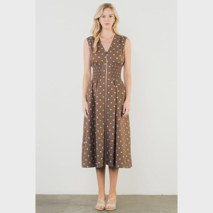 A woman stands against a plain background wearing the Zertz SLVLESS POLKA DOT MIDI DRESS, featuring brown polka dots, a V-neckline, and a fitted waist for an elegant look.