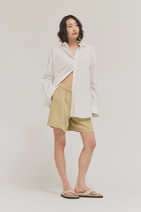 A woman with shoulder-length wavy hair poses casually against a plain background, wearing a loose white shirt, Zertz TENCEL SHORTS in light beige made from sustainable fabric, and beige sandals.