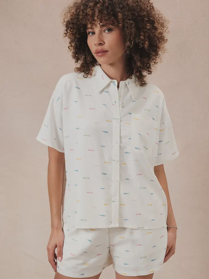 A woman with curly hair wears the Zertz PRINTED FISH SHIRT and matching shorts, both in white with a subtle colorful print, standing against a beige background that creates relaxed coastal vibes.