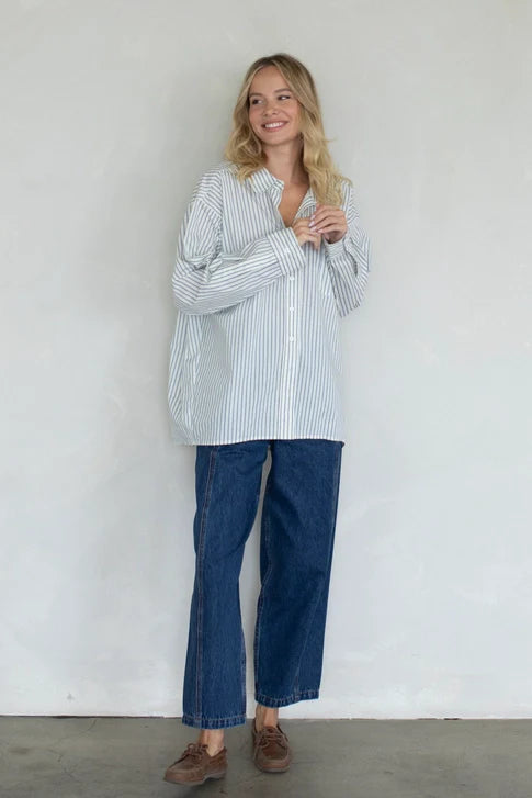 A person with long blonde hair wears the Zertz STRIPED OVER SIZED BUTTON UP TOP, paired with relaxed blue jeans and brown shoes, standing and smiling against a plain light-colored wall.