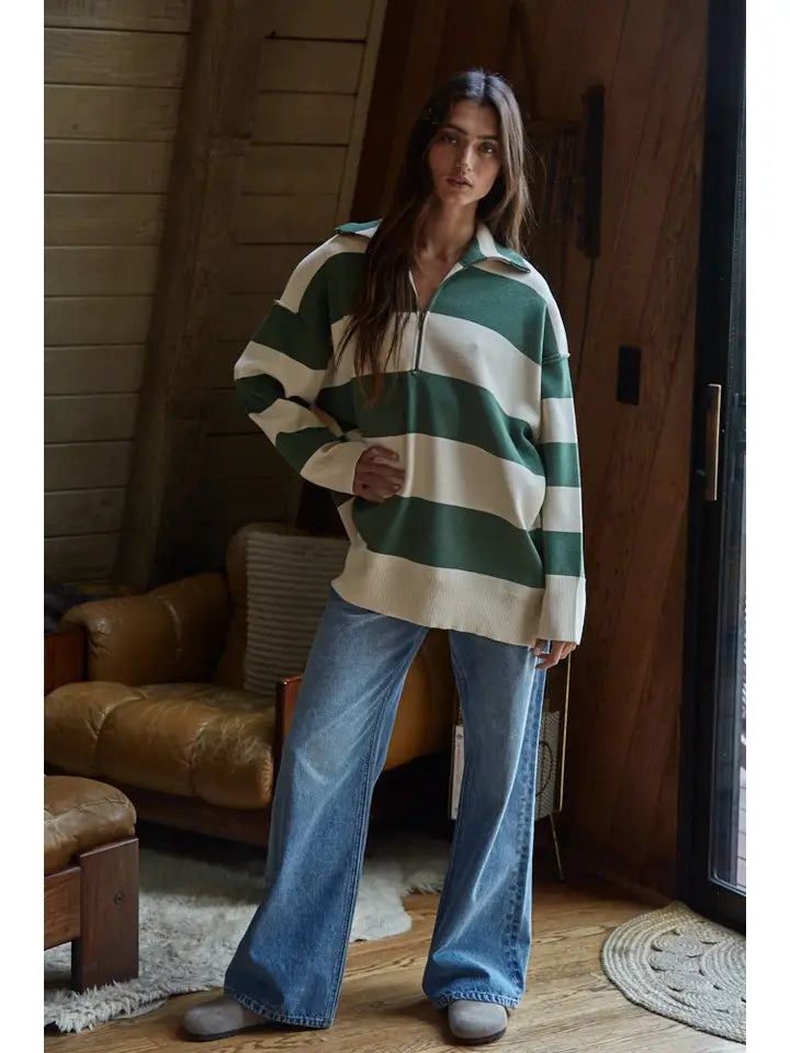 A woman stands indoors in a cozy, wood-paneled room, wearing the Zertz FONTAINE HALF ZIP SWEATER with green and white stripes, wide-leg blue jeans, and slippers. Natural light streams in onto a brown leather chair as she relaxes.