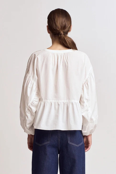 A woman with brown hair in a low ponytail is seen from behind, wearing the Zertz BALOON SLEEVE BLOUSE paired with dark blue jeans, standing against a plain light background.