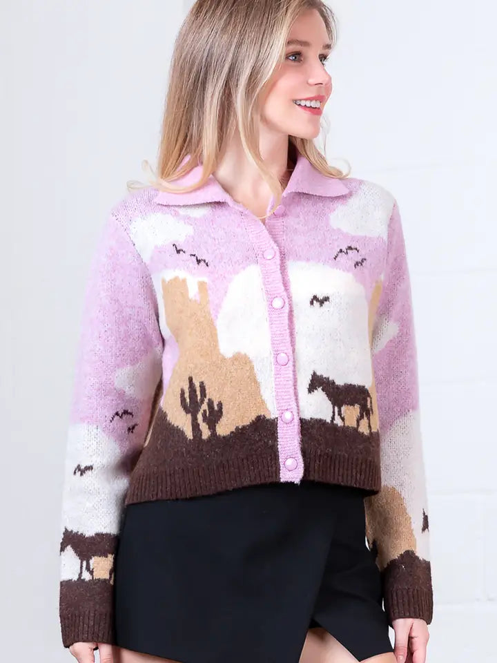 A woman with long blonde hair wears the Zertz JESSIE HORSE KNIT SWEATER in pink and brown, featuring a desert scene with cacti, horses, and birds. She smiles while looking to the side, paired with a black skirt.