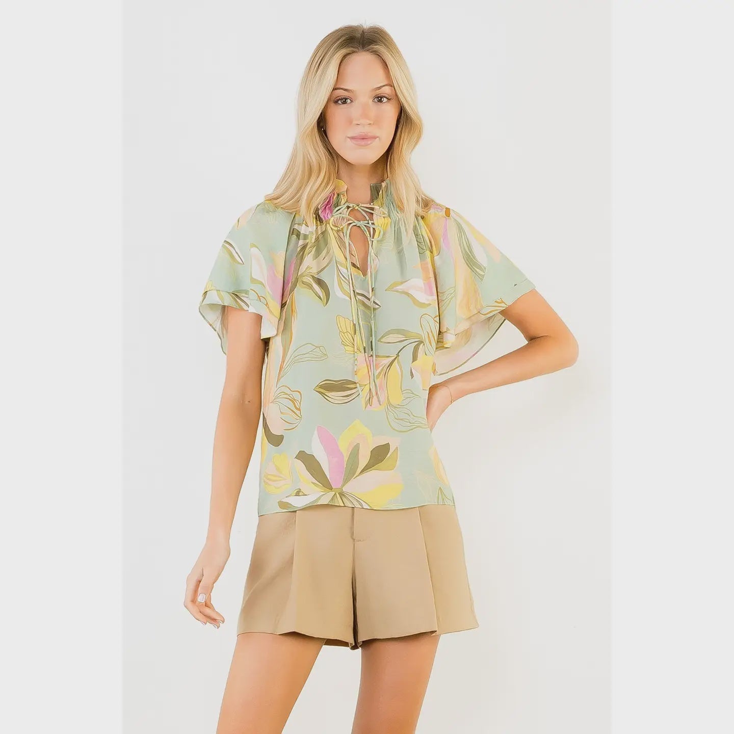 A woman models the Zertz FLOWY FLOWER TOP WITH TIE FRONT in light green, featuring short flutter sleeves and lightweight fabric, paired with beige shorts. She poses against a plain white background with one hand on her hip.
