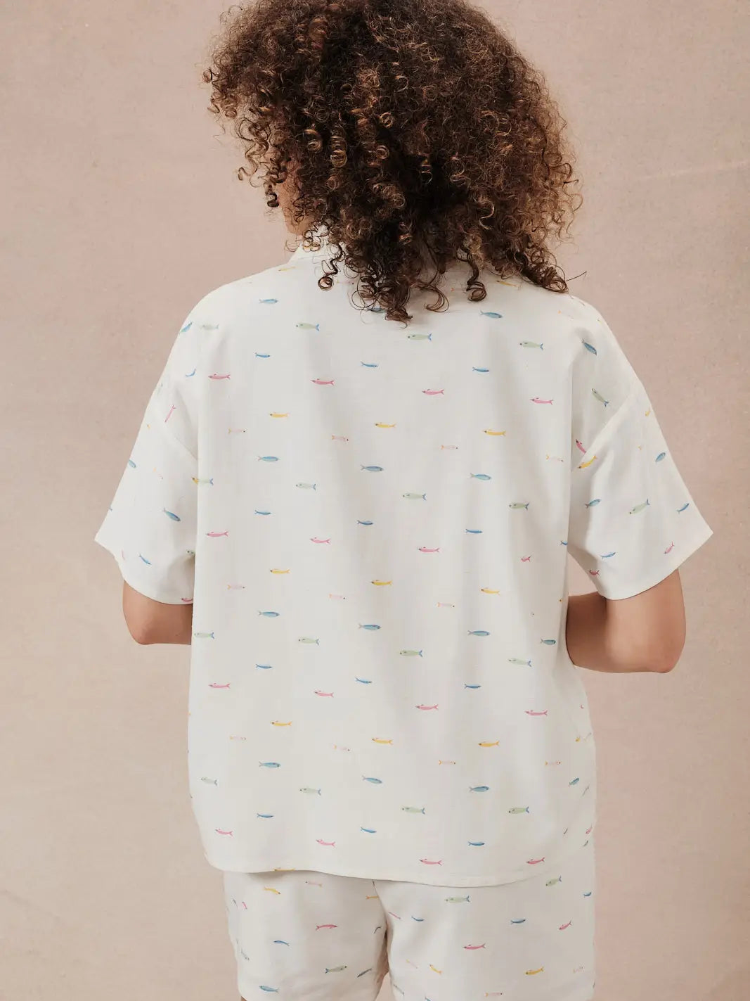 A person with curly hair faces away from the camera, wearing Zertz’s FISH PRINTED SHORTS and a loose white short-sleeve shirt—an ideal summer outfit featuring colorful fish patterns, set against a plain, light background.
