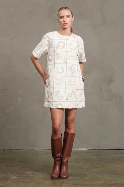 A woman stands against a plain gray wall, wearing the Zertz WESTERN EMB MINI DRESS with brown graphic designs and knee-high brown boots. Her hair is pulled back, and her hands rest in her pockets.