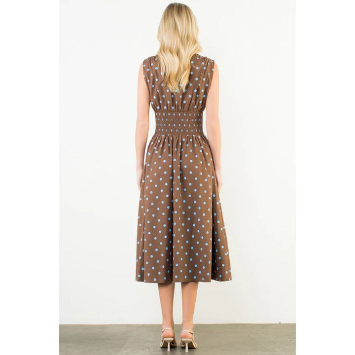 A woman with long blonde hair wears the Zertz SLVLESS POLKA DOT MIDI DRESS, featuring light blue dots on brown fabric and a gathered waist. She pairs it with beige heeled sandals, standing on a gray floor against a white background.