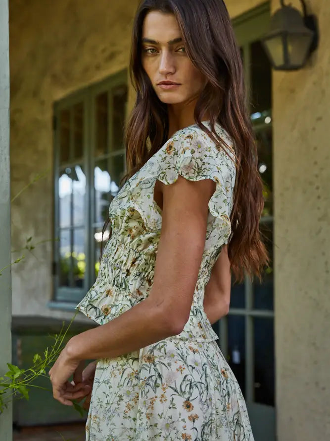 A woman with long brown hair wears the SEREIA FLORAL TOP by Zertz, standing outdoors near a window and green door, looking over her shoulder with a neutral expression.