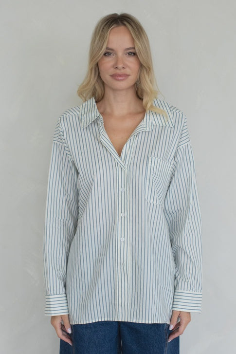 A woman with blonde hair stands against a light background, wearing the Zertz STRIPED OVER SIZED BUTTON UP TOP paired with blue jeans. She faces the camera, her expression neutral and composed.