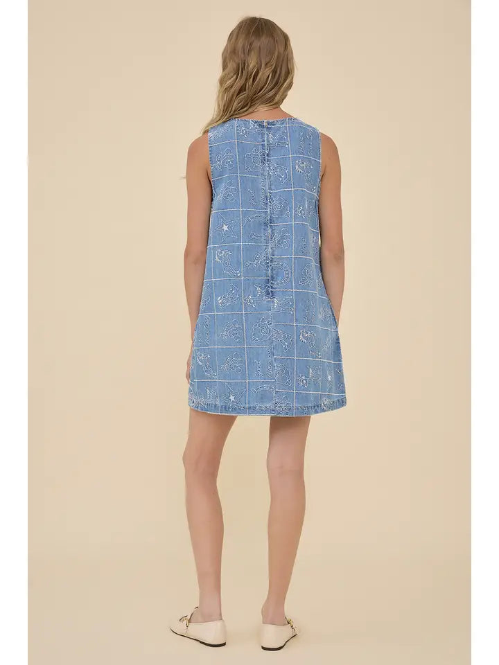 A woman with wavy hair, seen from behind, wears the Zertz EMBROIDERED DENIM WESTERN DRESS featuring a white grid and floral pattern. She stands against a beige background, embracing a cowgirl vibe with classic white slip-on shoes.