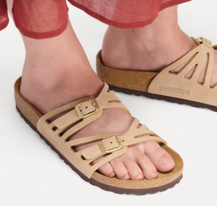 A person in a flowing red skirt stands on a white surface, wearing Zertz GRANADA LENB SANDCASTLE R sandals with nubuck leather, two gold buckles, and brown soles for a natural look.