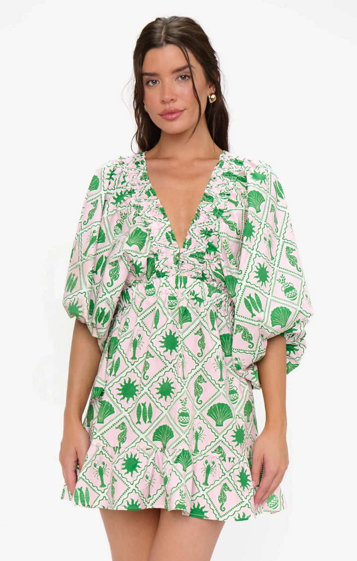 A woman wears the Zertz HADLEY MINI DRESS, featuring a deep V-neckline, puff sleeves, and a geometric green and white plant-and-sun motif resembling a coastal collage. She stands against a plain white background.