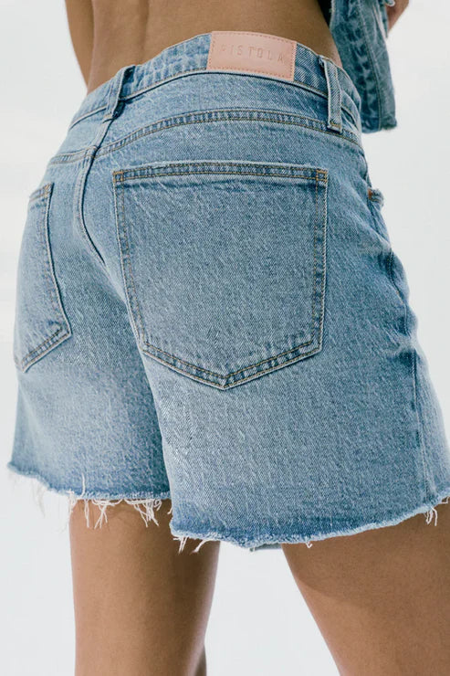Close-up of a person wearing Zertz KENNEDY RELAXED MID RISE CUT OFF SHORT in sun-washed blue comfort stretch denim, with frayed hems and a pink waistband label. Shot from behind against a plain background.