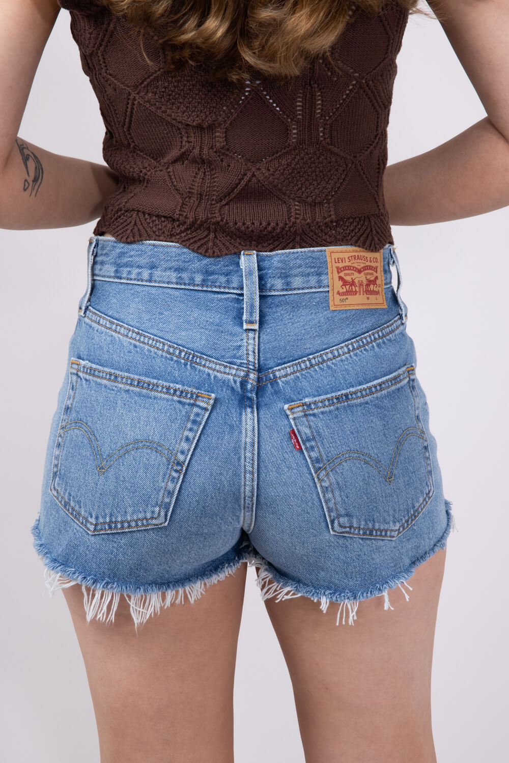 A person with long brown hair wears a waist-defining brown sleeveless knit top and leg-lengthening blue Zertz 501 ORIGINAL SHORT, shown from the back against a light plain background. A tattoo appears on their upper right arm.