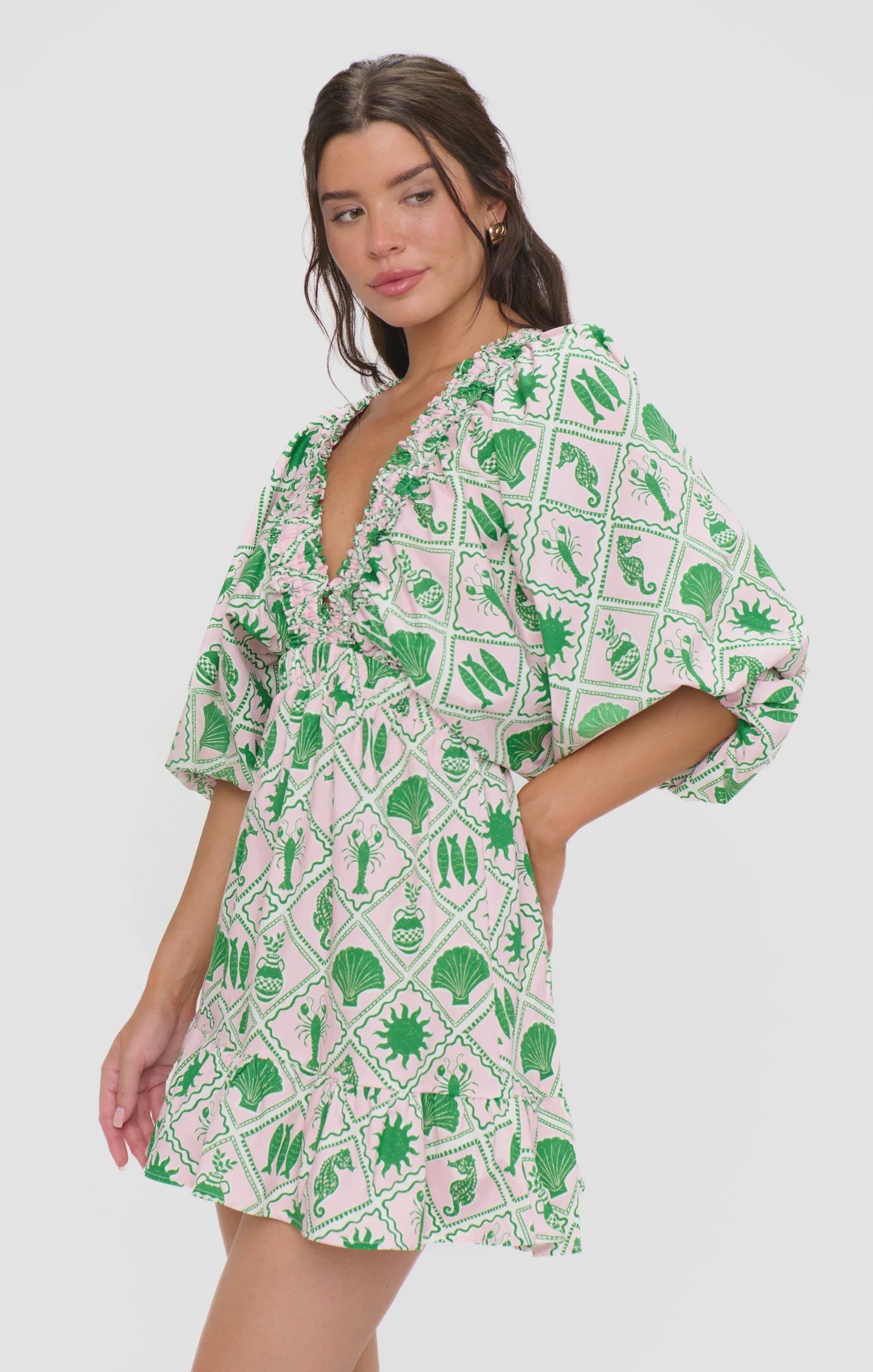 A woman models the Zertz HADLEY MINI DRESS, showcasing a coastal collage print with green and white geometric patterns, ruffled trim, deep V-neck, and puff sleeves against a plain white background.