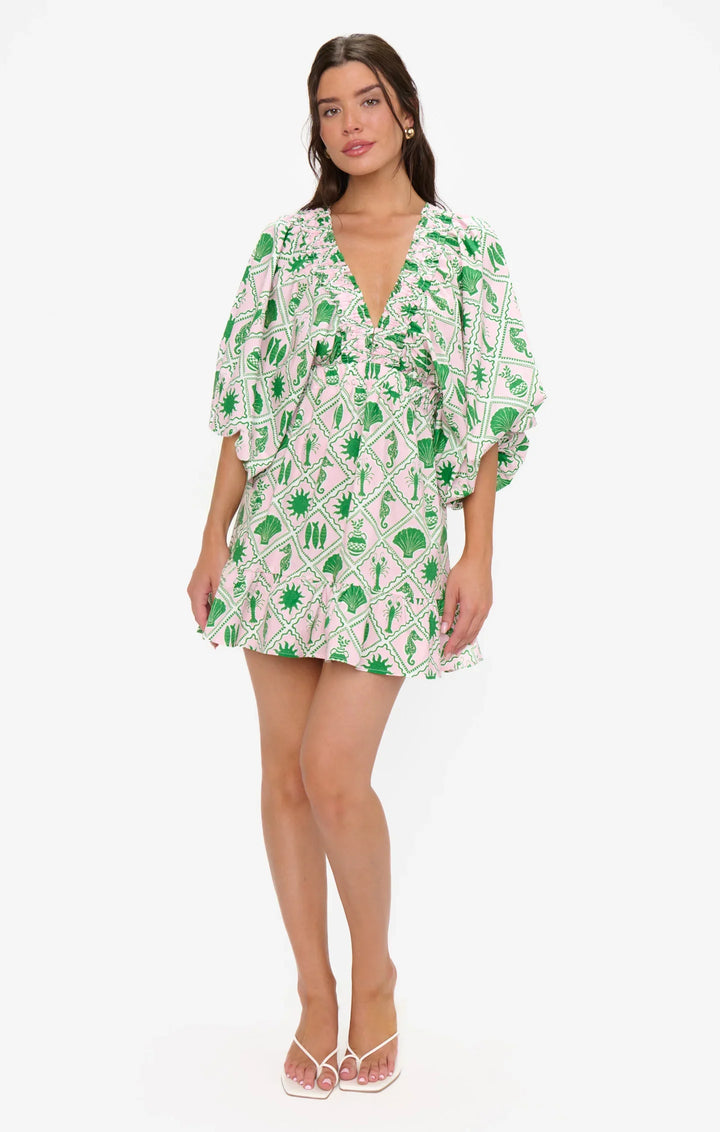A woman poses against a white background in the Zertz HADLEY MINI DRESS, a short, flowy mini dress with puff sleeves, green and white geometric floral print, deep V-neck, and ruffled trim, styled with white sandals.