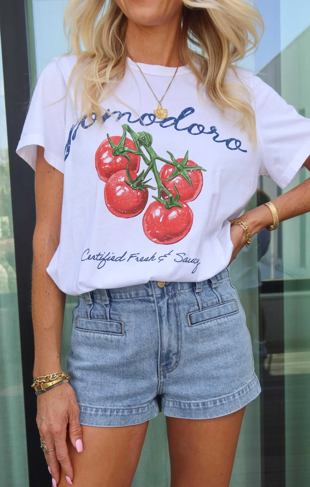 A woman wears the Zertz TRAVIS TEE, featuring a bold tomato graphic and "pomodoro," paired with high-waisted denim shorts, gold bracelets, and necklaces, posing with one hand on her hip in front of a glass door.