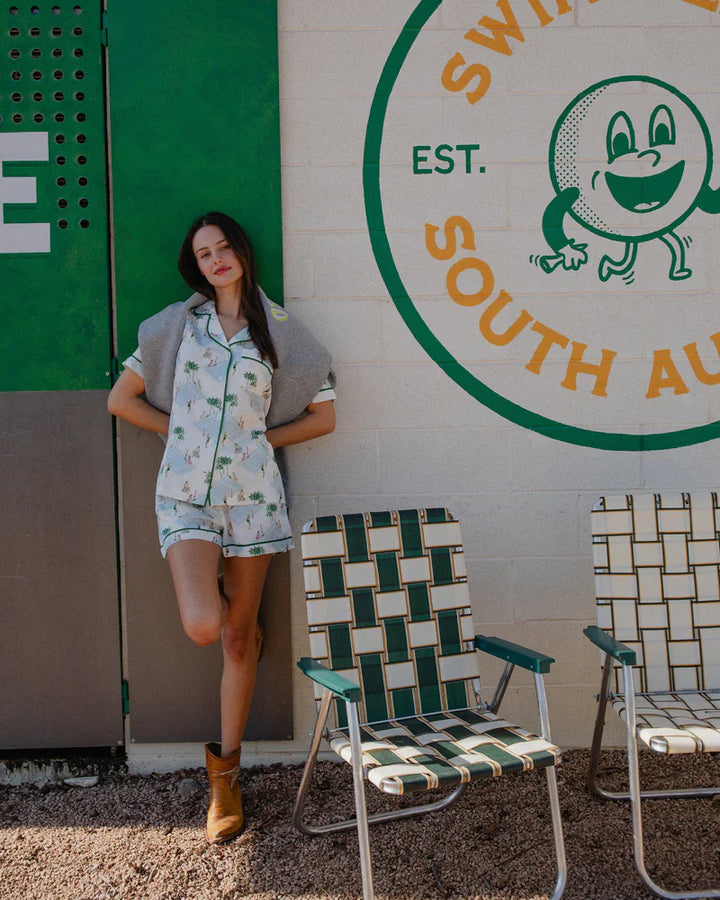 Wearing the Zertz PALM COURTS PAJAMA SHORT SET, a woman in boots leans against a mural wall that reads "South Aus" with a bright orange illustration. Two woven lawn chairs sit on gravel nearby.