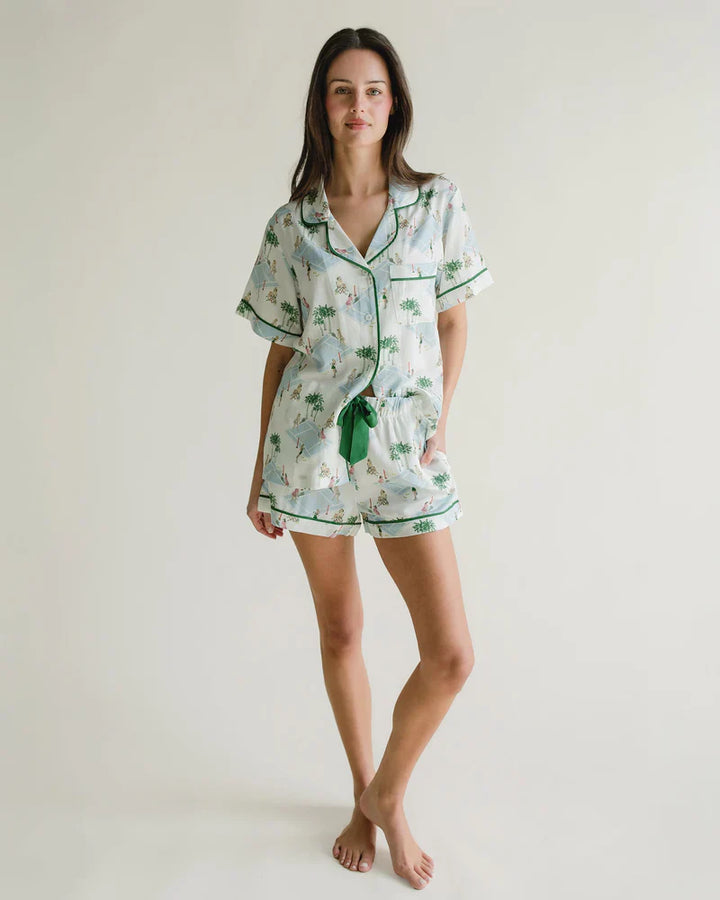 A woman stands barefoot against a plain background, wearing the Zertz PALM COURTS PAJAMA SHORT SET with a green palm tree print, green trim, and an elastic waist. She has her hands in her shorts pockets and faces the camera.