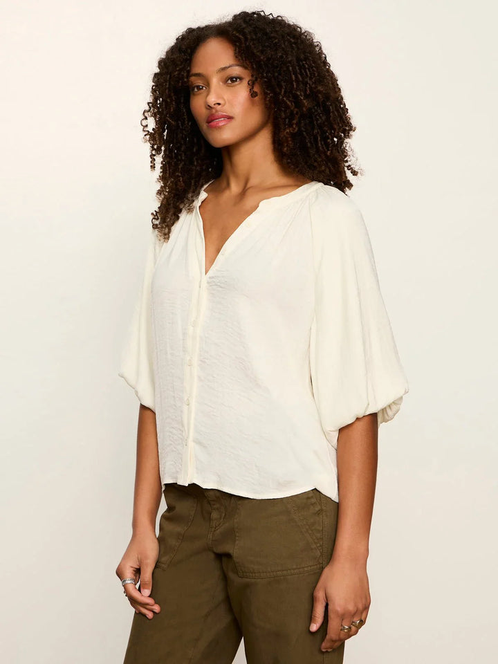 A woman with curly hair models the Zertz PLENTY OF LOVE BUBBLE BLOUSE, a relaxed fit off-white top with a split neck and three-quarter sleeves, paired with olive green pants against a plain light background.