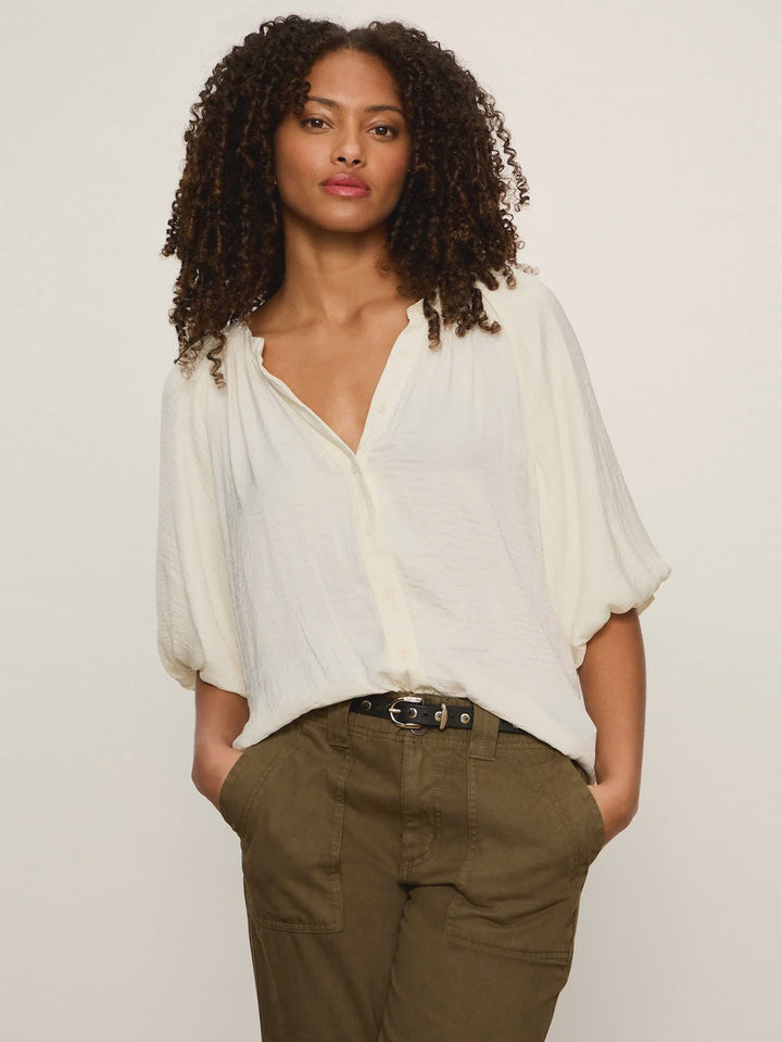 A woman with curly hair wears the Zertz PLENTY OF LOVE BUBBLE BLOUSE, a relaxed fit with bubble short sleeves, tucked into olive green pants. She stands against a light background with one hand in her pocket.
