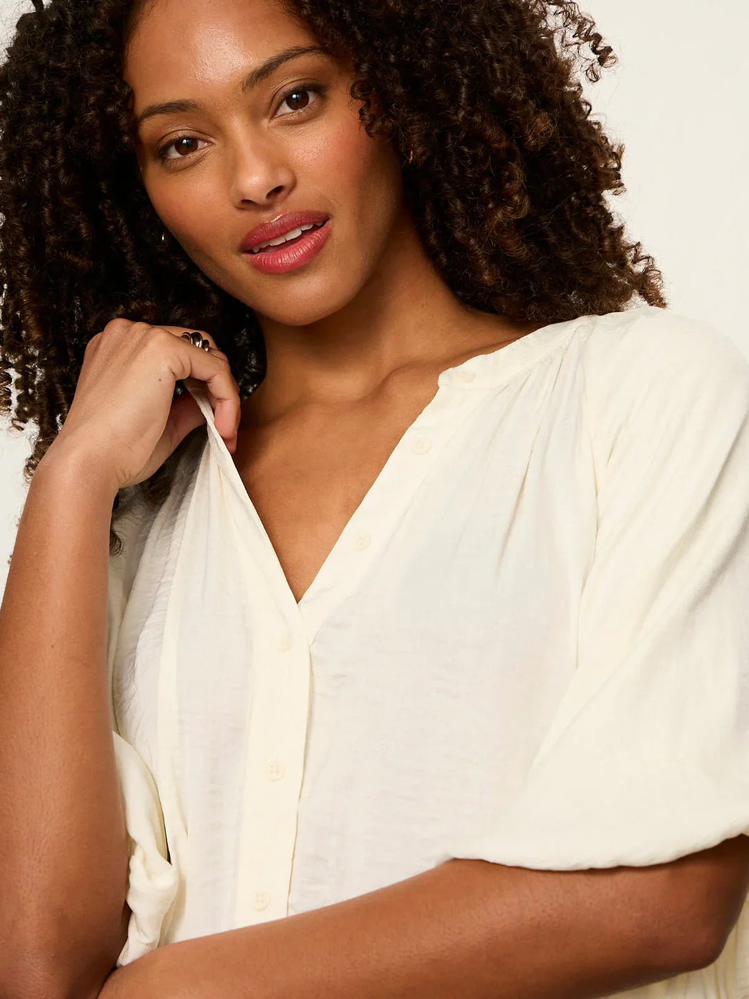 A woman with curly hair and natural makeup models the Zertz PLENTY OF LOVE BUBBLE BLOUSE, a relaxed fit cream split neck top, holding the collar and looking confidently at the camera against a plain background.
