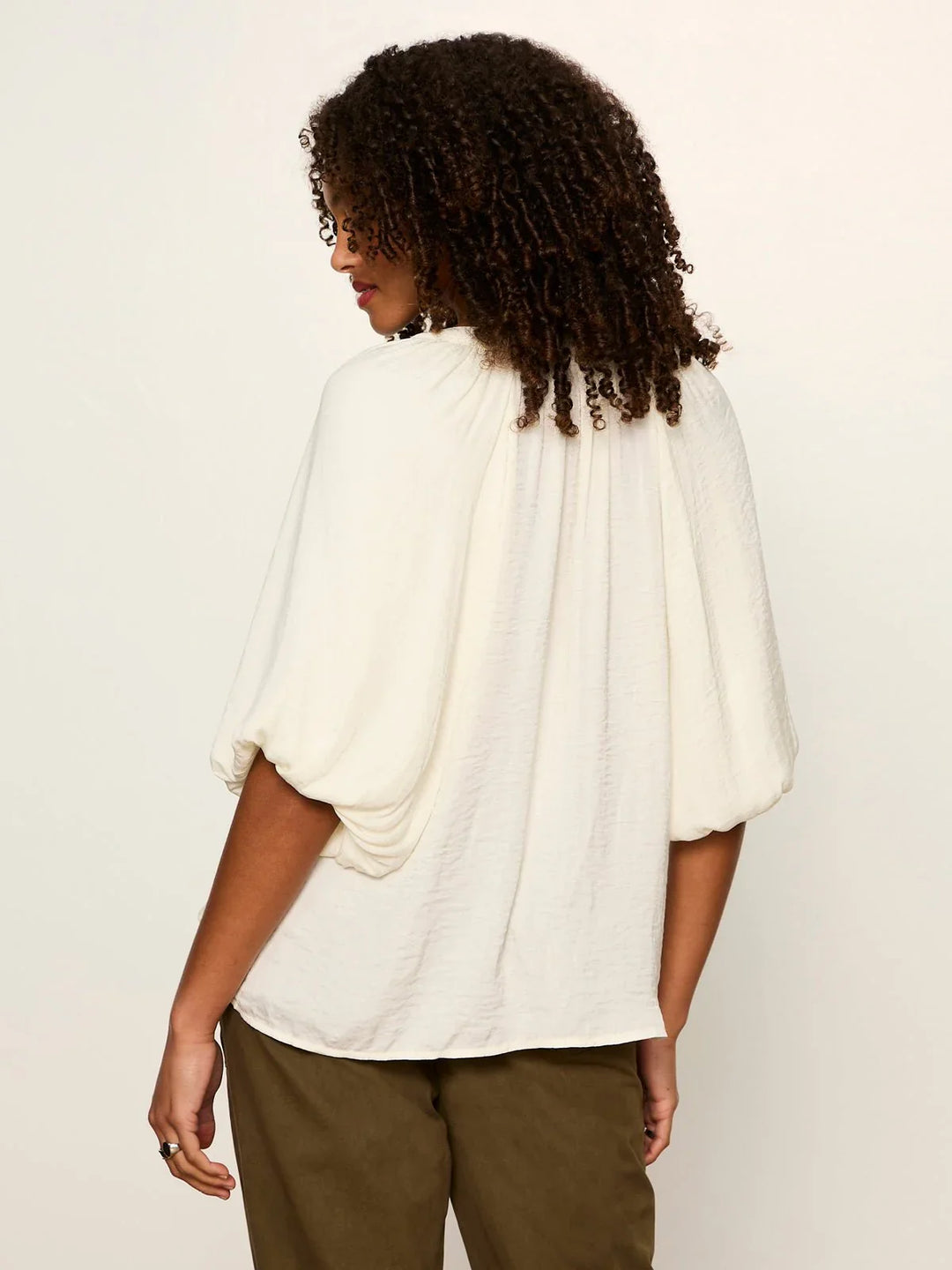 A person with curly hair stands with their back to the camera, wearing the off-white PLENTY OF LOVE BUBBLE BLOUSE by Zertz and olive green pants against a plain, light background.