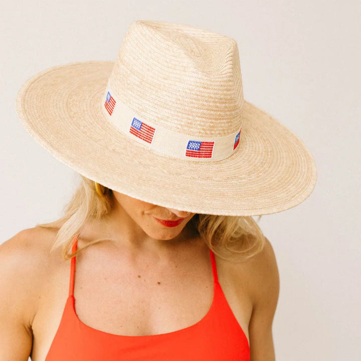 A woman wears the Zertz AMERICA PALM HAT, a sustainable ivory hat with a wide brim, small American flags around the crown, and UPF sun protection, paired with a red sleeveless top against a plain background.
