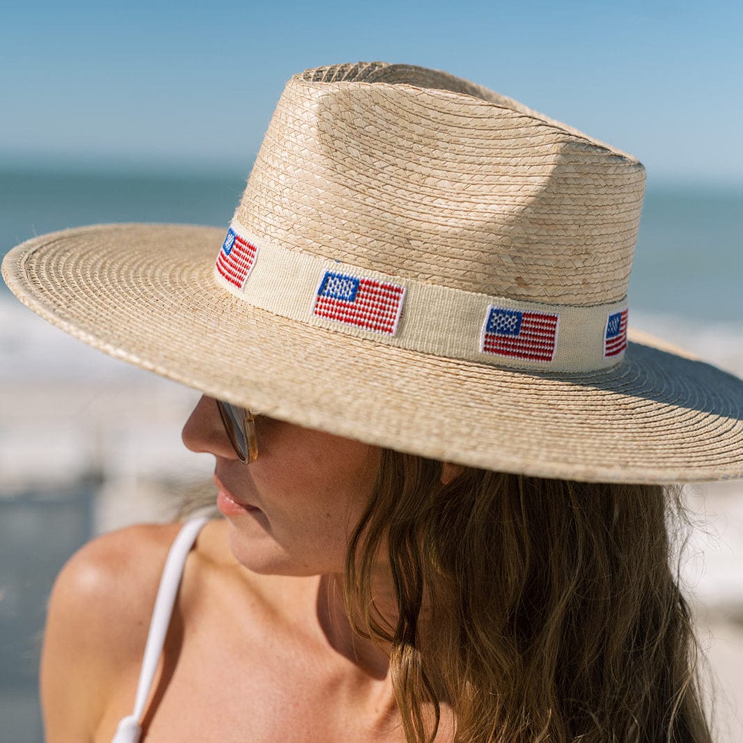 A woman in sunglasses and a Zertz AMERICA PALM HAT—made from sustainable materials with UPF sun protection—stands on a beach decorated with small American flags, the ocean and sky in the background.