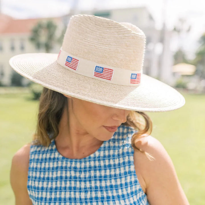 A woman with wavy brown hair wears the Zertz AMERICA PALM HAT, featuring an ivory shade, UPF protection, and American flag band. She pairs it with a blue and white checkered sleeveless top while outdoors on a sunny day.
