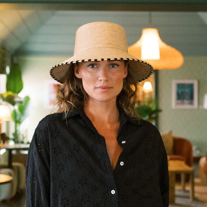 A woman wears a black button-up shirt and the Zertz JAKELINE BUCKET HAT, featuring a wide brim and black trim, as she stands indoors in a stylish, plant-filled room with warm lighting.