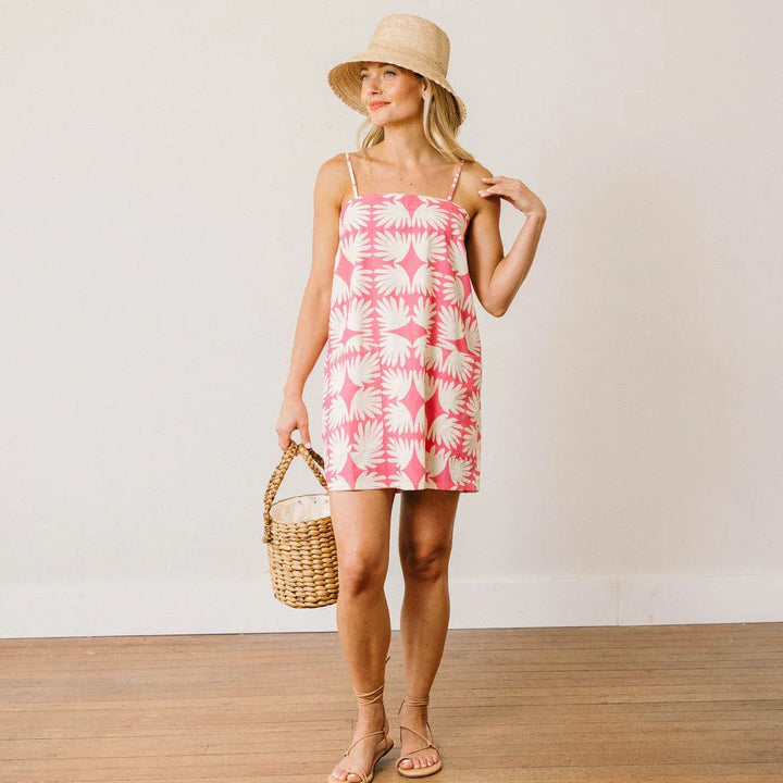 A woman wears the Zertz MILA BUCKET HAT, a pink and white patterned sleeveless dress, and sandals indoors while holding a woven basket purse, smiling and looking to the side.