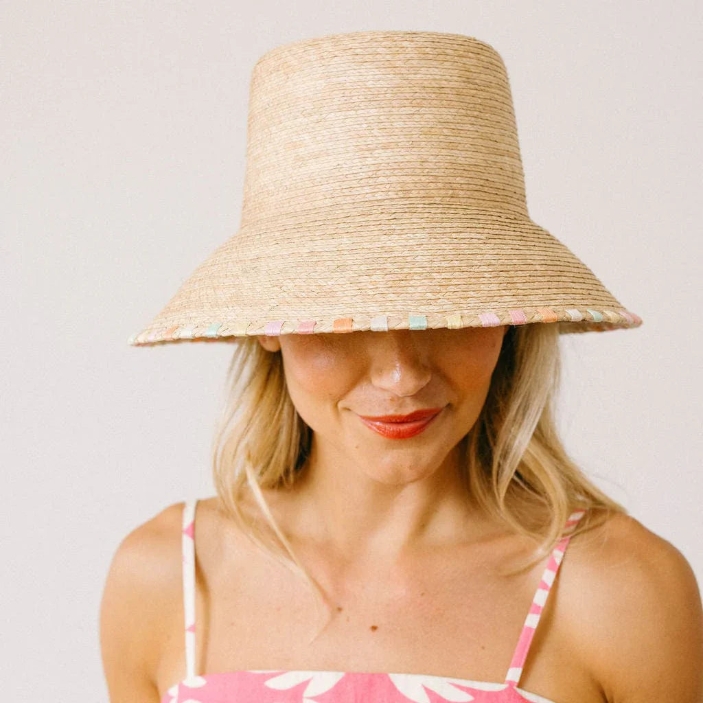 A woman with blond hair and red lipstick wears the Zertz MILA BUCKET HAT—a wide, woven straw UPF 50+ hat with a pastel-trimmed brim—partially covering her eyes, paired with a pink patterned, thin-strapped top.