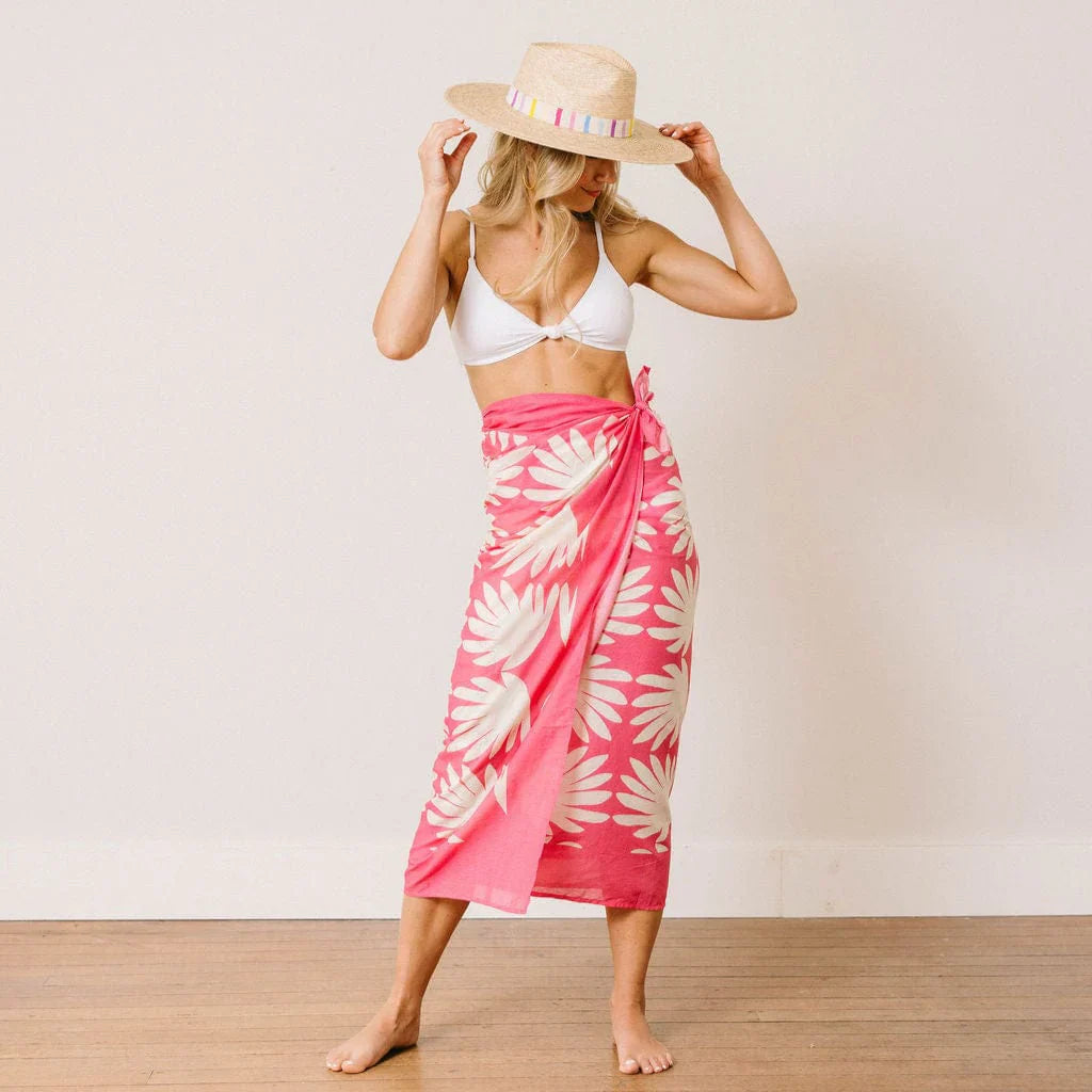 A woman stands barefoot on a wooden floor against a plain white wall, looking down and adjusting her Zertz VALENTINA PALM HAT while wearing a white bikini top and pink and white floral sarong.