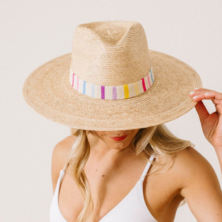 A woman in a white swimsuit wears the Zertz VALENTINA PALM HAT—a wide-brimmed, fair trade hat featuring a colorful striped band—holding the brim and looking down.