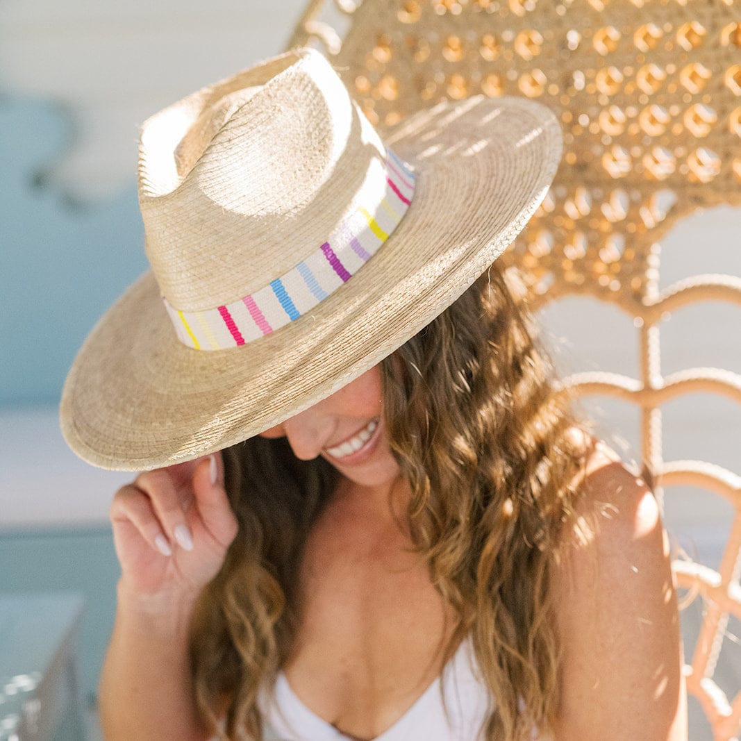 A woman with wavy brown hair smiles on a woven chair, tipping the Zertz VALENTINA PALM HAT—featuring a colorful striped band and UPF protection—over her face as sunlight brightens the relaxed, summery scene.